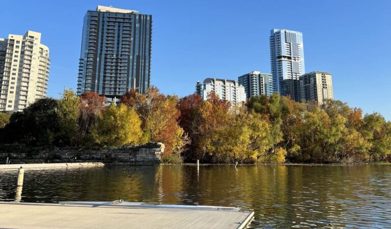 A lake in Austin, Texas, with a a dock in the foreground, trees filled with orange and yellow leaves on the edge of the lake, and a skyline of skyscrapers in the background
