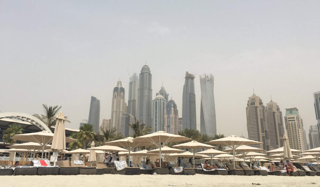 A row of lounge chairs and umbrellas on a beach with the tall skyscrapers of Dubai in the background