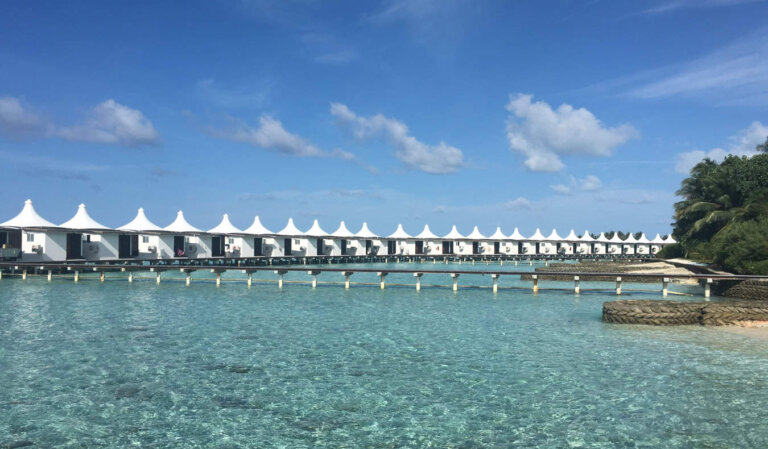 A row of white overwater bungalows connected by a long wooden walkway over the crystal clear waters of the Maldives