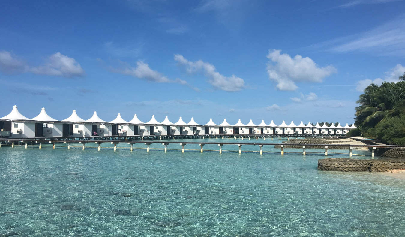 A row of white overwater bungalows connected by a long wooden walkway over the crystal clear waters of the Maldives