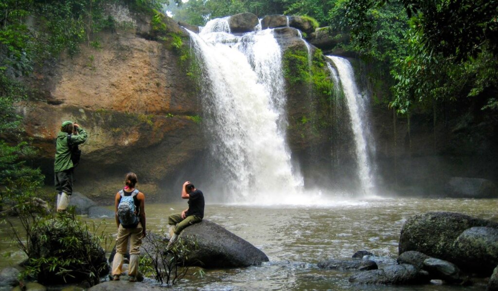 three people sitting on rocks near a waterfall in the lush jungle of Khao Yai National Park in Thailand