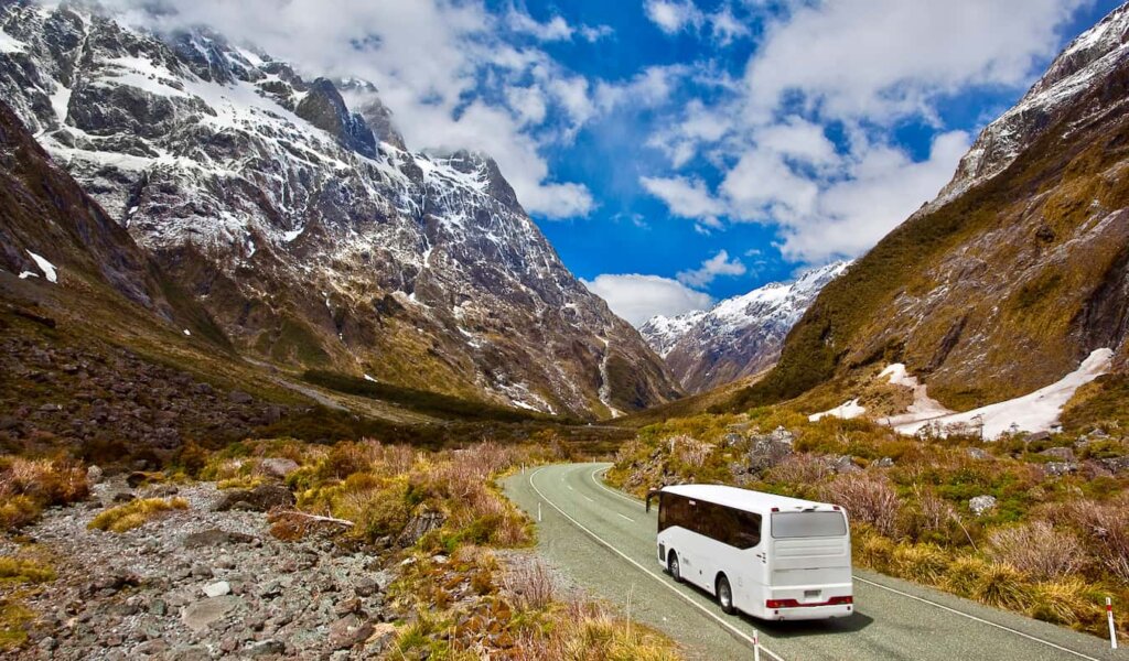 A bus driving down a winding road through tall snowcapped mountains in New Zealand