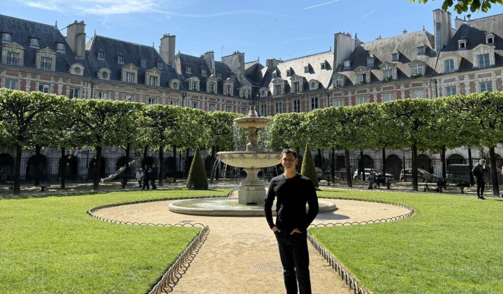 Nomadic Matt standing in the Place de Vosages, a large enclosed square surrounded by buildings, with a fountain in the middle, in Paris, France