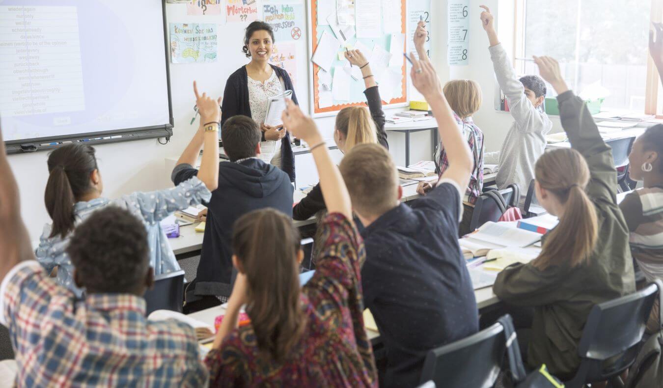 A teacher stands in front of a white board looking out at a class with many hands raised