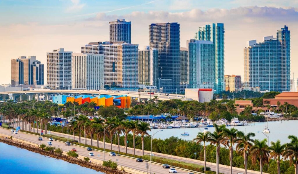 The downtown skyline of Miami at sunset, with skyscrapers in the background and a palm-tree-lined waterfront boulevard in the foreground